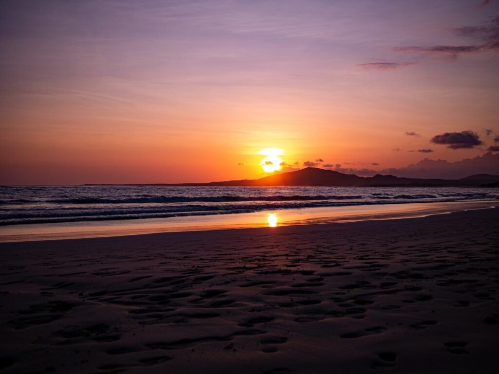 the sun is setting over the ocean with footprints in the sand